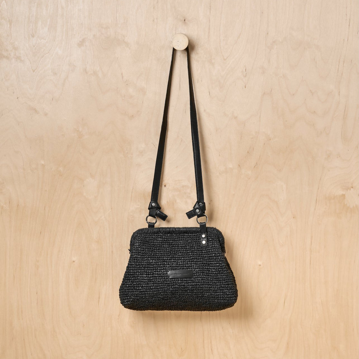 Black woven shoulder bag hanging on a light wood wall.