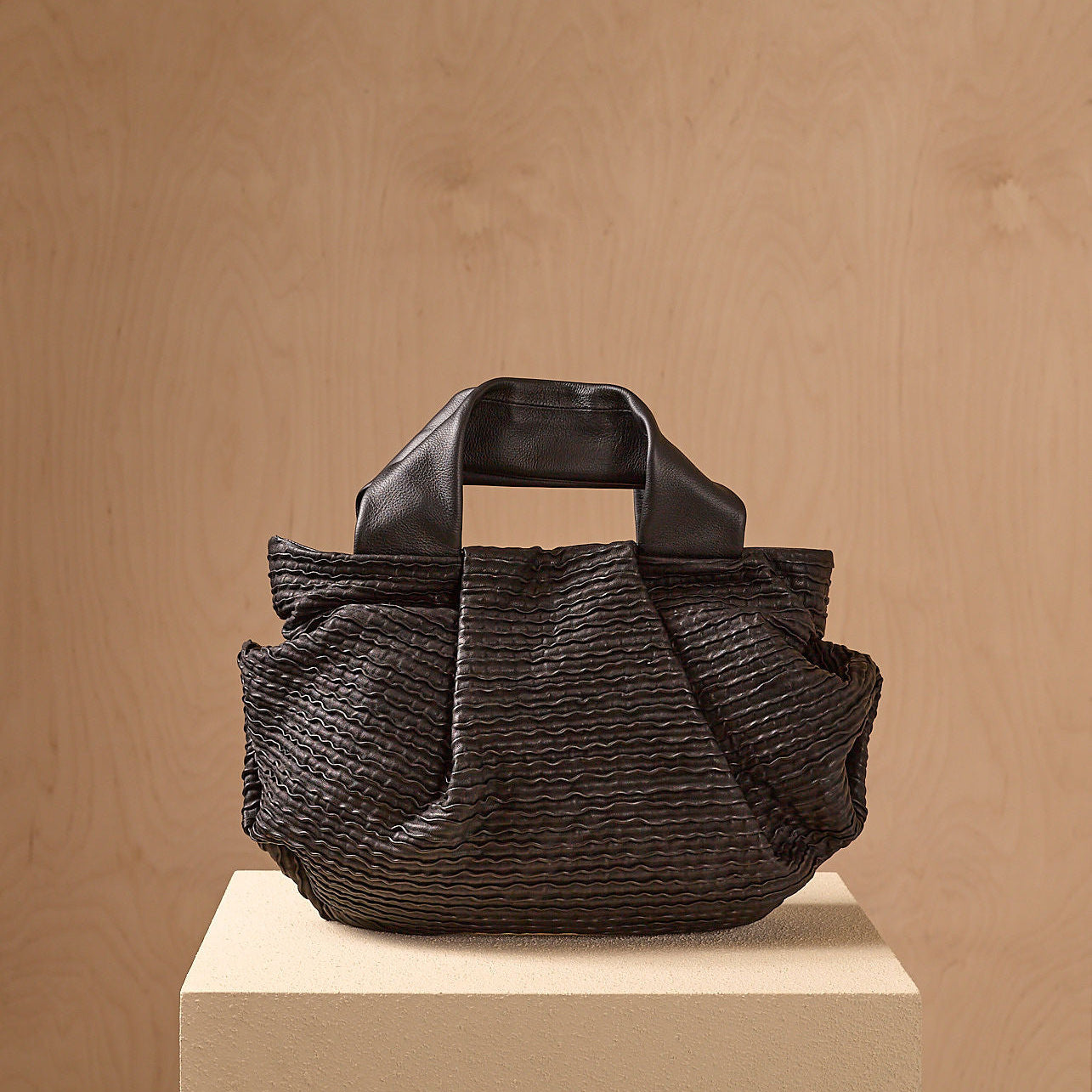 A black wavy textured handbag with leather handles placed on a wooden pedestal against a wooden background.