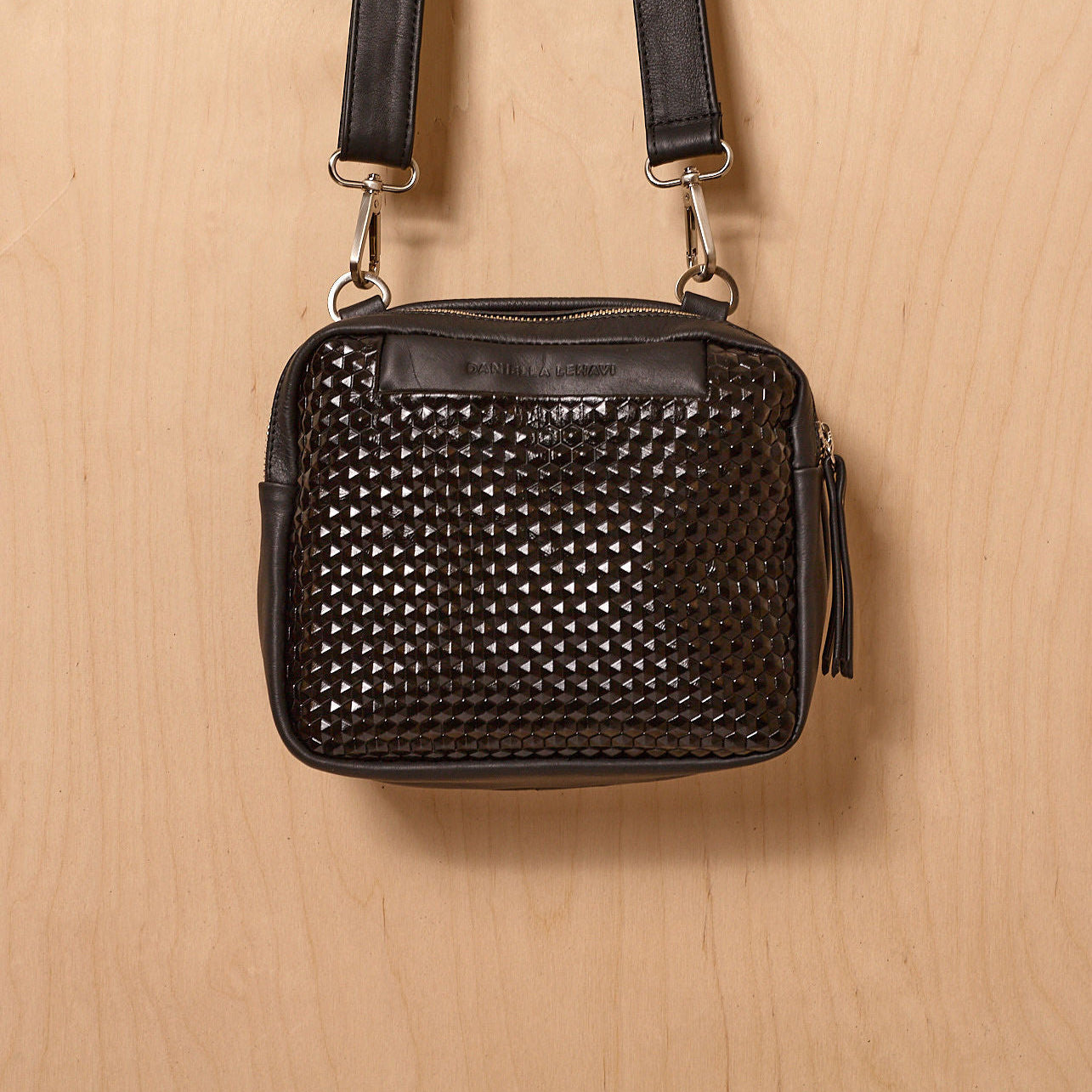 תיק צד שחור של דניאלה להבי עם רצועת כתף ודוגמת פירמידות מבריקה, על רקע עץ בהיר.