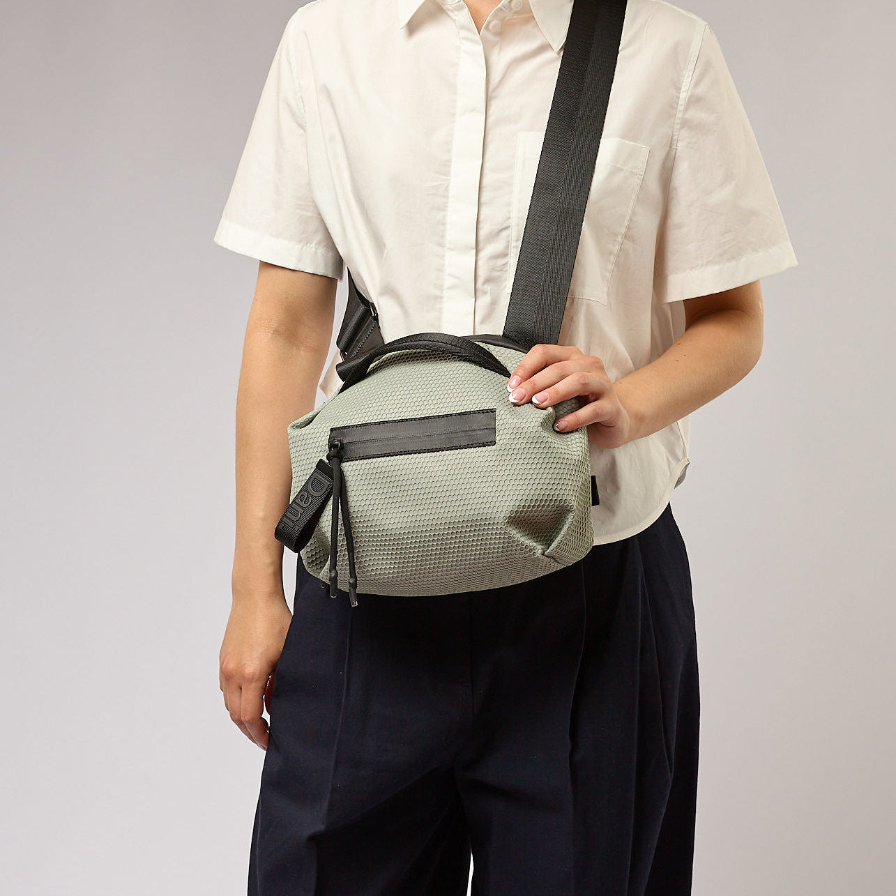 אישה עונדת תיק צד ירוק בהיר בדוגמת כוורת עם רצועה שחורה, על רקע אפור.