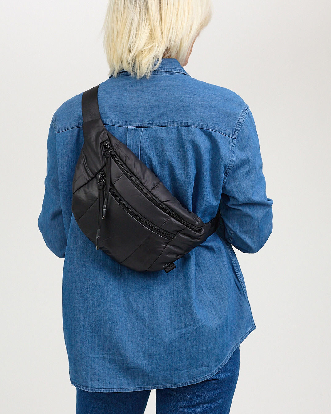 A person wearing a blue denim jacket is shown from the back, carrying a black quilted belt bag with zippered compartments and an adjustable strap.