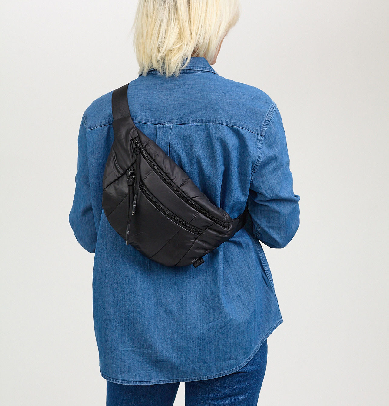 A person wearing a blue denim jacket is shown from the back, carrying a black quilted belt bag with zippered compartments and an adjustable strap.