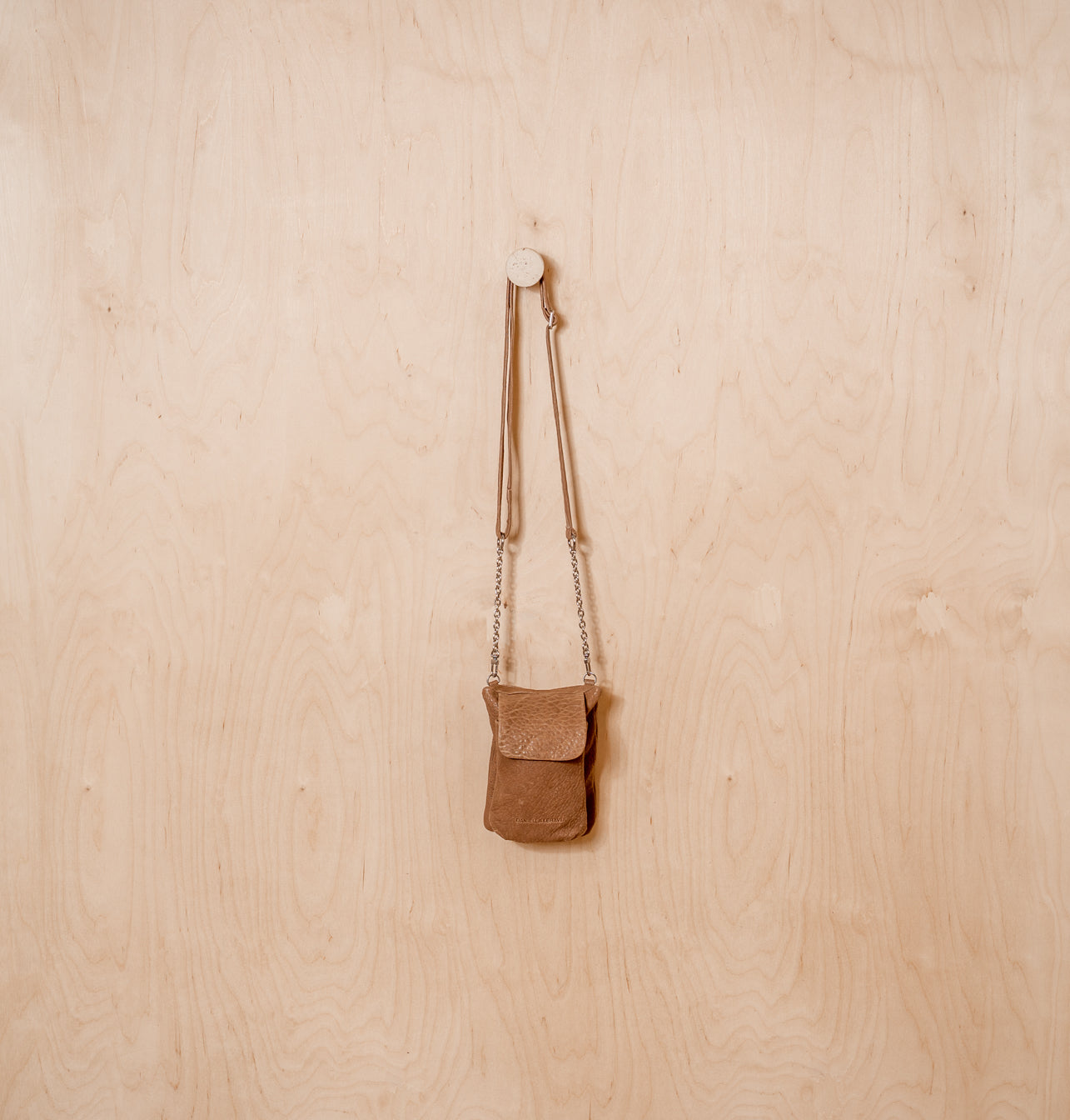 תיק צד קטן חום עם רצועת שרשרת תלוי על קיר עץ בהיר.
