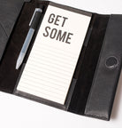 מארגן עור שחור פתוח, עם פנקס 'GET SOME', עט שקוף וכפתור סגירה.